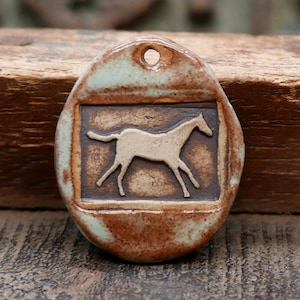 May include: An oval ceramic pendant featuring a raised cream-colored horse silhouette centered within a dark brown rectangular frame. The pendant has a mottled light blue and brown glazed finish and includes a small circular hole at the top.