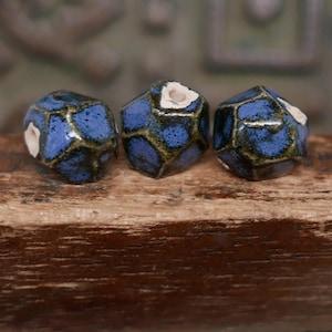 May include: Three blue ceramic beads with a geometric design. The beads have a dark blue base color with lighter blue and dark green accents. Each bead has a unique shape and a small white area. They are displayed on a wooden surface.