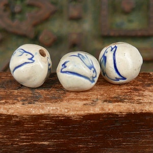 May include: Three off-white ceramic beads with blue painted designs. One bead has a hole. The designs include a rabbit and a bird. The beads are on a wooden surface. These beads are suitable for jewelry making or other craft projects.