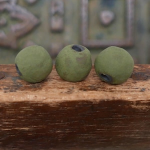 May include: Three olive-green, spherical objects with dark openings are arranged on a weathered wooden surface. The objects have a textured appearance and are evenly spaced, suggesting a collection or display of decorative items.