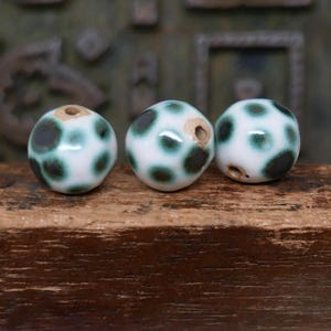 May include: Three white ceramic beads with a speckled pattern of dark green spots. Each bead has a hole running through the center. The beads are displayed on a wooden surface.