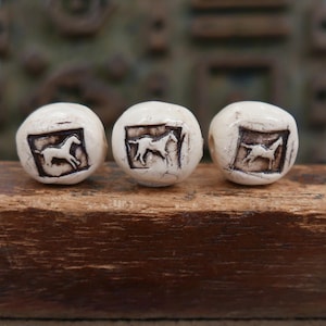 May include: Three off-white, rounded beads with a cracked, textured surface. Each bead features a square, dark brown recessed panel containing a stamped, white silhouette of a horse in a different pose, showing a running or standing animal.