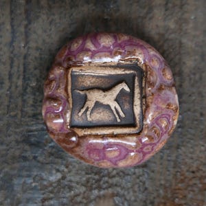 May include: A ceramic horse-themed button with a square inset featuring a horse silhouette. The button has a rounded shape with a textured, patterned border in shades of purple and brown. The horse is tan against a dark brown background.