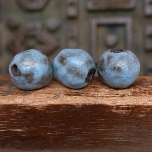 May include: Three ceramic beads with a light blue glaze and dark gray speckling. Each bead has a hole and sits on a wooden surface. The beads are round and have a slightly irregular shape, resembling blueberries.