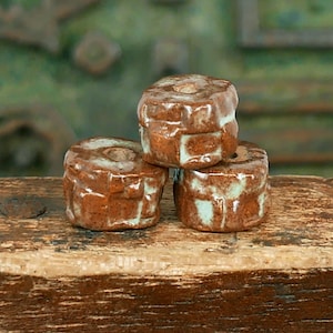 May include: Three brown ceramic beads with white glaze accents. The beads are stacked on top of each other on a wooden surface.
