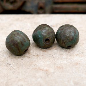 May include: Three weathered, round beads with a dark, mottled appearance. The beads have a greenish-brown patina and a hole drilled through the center. The beads are likely made of metal and are suitable for jewelry making or other crafts.