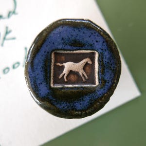 May include: A round, ceramic bottle stopper with a dark blue glaze. The center features a square with a brown background and a white horse silhouette. The stopper is likely handmade and has a rustic, artisanal feel.