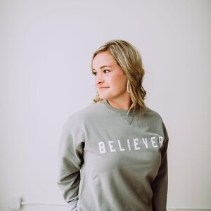 May include: A woman wearing a gray sweatshirt with the word "BELIEVER" printed in white on the front. She is standing in front of a white wall.