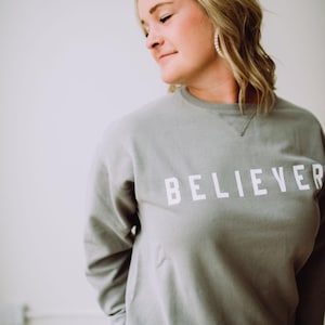 May include: A woman wearing a gray sweatshirt with the word "BELIEVER" printed in white on the front.