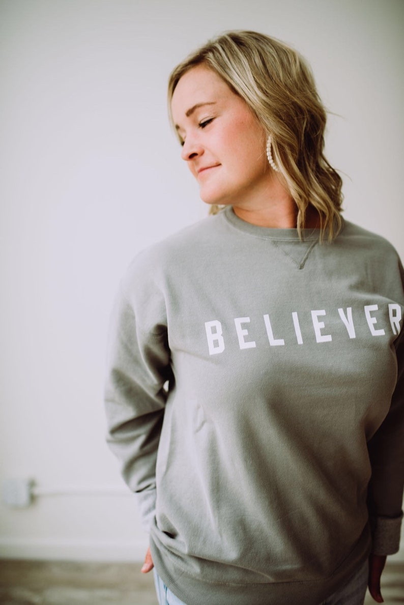 May include: A woman wearing a gray sweatshirt with the word "BELIEVER" printed in white on the front.