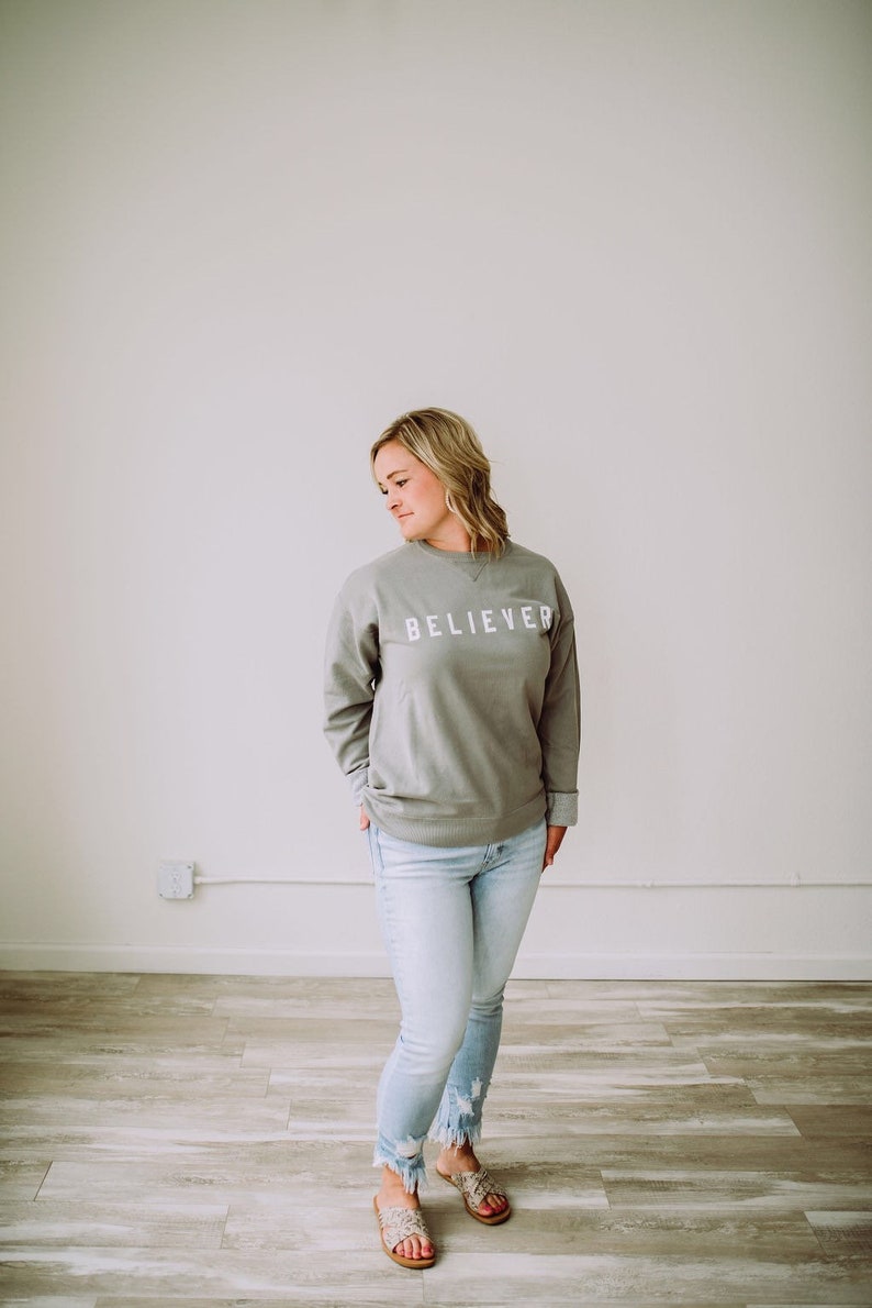 May include: A woman wearing a light gray sweatshirt with the word "BELIEVER" printed on the front. She is wearing light blue jeans and brown sandals.