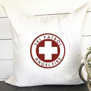 Può includere: Cuscino bianco con un design a cerchio rosso che dice "Ski Patrol Angel Fire" con una croce rossa al centro.