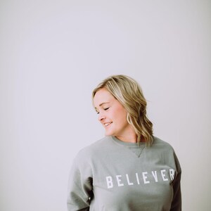 May include: A woman wearing a gray sweatshirt with the word "BELIEVER" printed in white on the front. She is standing in front of a white wall.