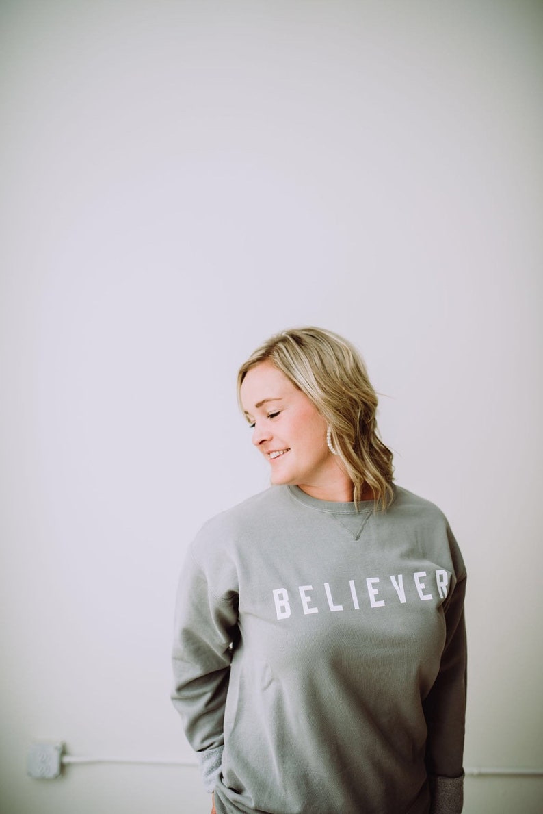 May include: A woman wearing a gray sweatshirt with the word "BELIEVER" printed in white on the front. She is standing in front of a white wall.