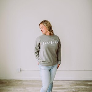 May include: A woman wearing a light gray sweatshirt with the word "BELIEVER" printed on the front. She is wearing light blue jeans and brown sandals.