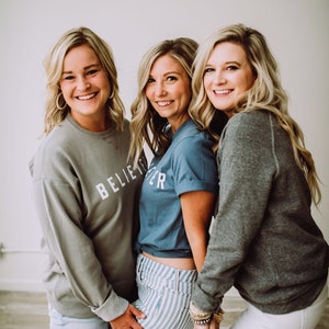May include: Three women are standing together, smiling. The woman in the middle is wearing a blue t-shirt with the text "Believe" on it. The woman on the left is wearing a gray sweatshirt with the text "Believe" on it. The woman on the right is wearing a gray sweatshirt.