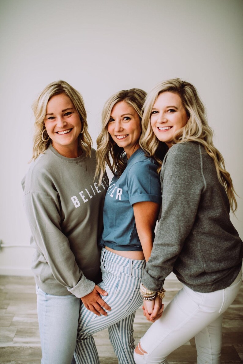 May include: Three women are standing together, smiling. The woman in the middle is wearing a blue t-shirt with the text "Believe" on it. The woman on the left is wearing a gray sweatshirt with the text "Believe" on it. The woman on the right is wearing a gray sweatshirt.