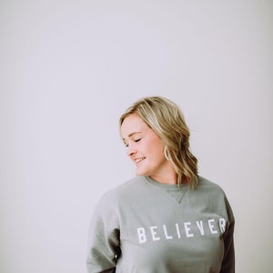 May include: A woman wearing a gray sweatshirt with the word "BELIEVER" printed in white on the front. She is standing in front of a white wall.