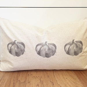 May include: A beige pillow with three gray pumpkins printed on it. The pumpkins are stylized and have a watercolor effect.