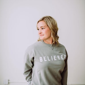 May include: A woman wearing a gray sweatshirt with the word "BELIEVER" printed in white on the front. She is standing in front of a white wall.
