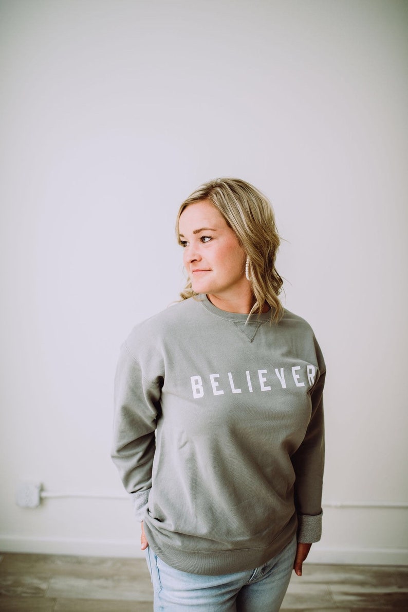 May include: A woman wearing a gray sweatshirt with the word "BELIEVER" printed in white on the front. She is standing in front of a white wall.