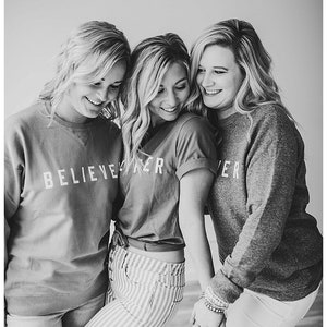 May include: Three women wearing matching shirts with the text "BELIEVE" on them. The woman in the middle is wearing striped pants and the women on either side are wearing jeans.