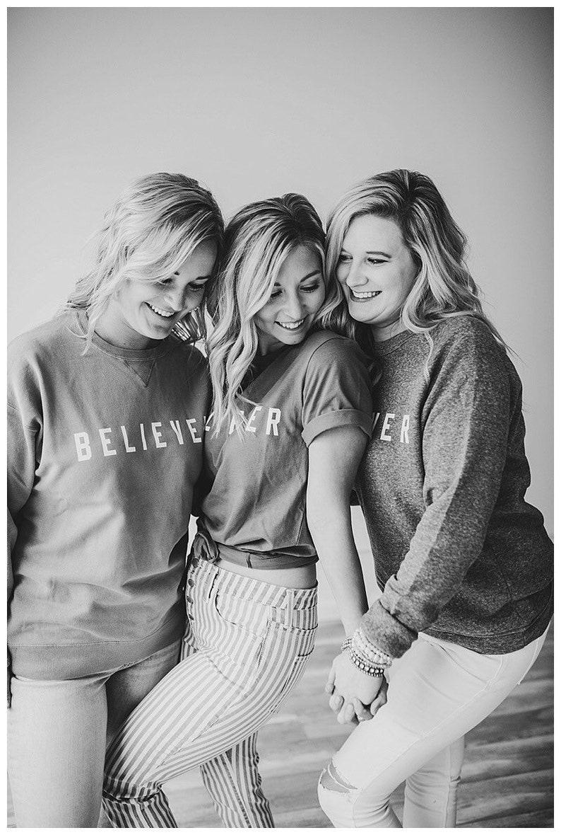 May include: Three women wearing matching shirts with the text "BELIEVE" on them. The woman in the middle is wearing striped pants and the women on either side are wearing jeans.