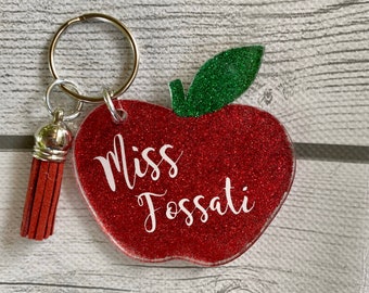May include: A red glitter apple-shaped keychain with a green glitter leaf. The keychain has the name "Miss Fossati" in white script. A red tassel hangs from the keychain ring.