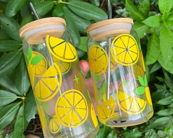 May include: Two clear glass tumblers with bamboo lids and metal straws. The tumblers have a lemon print design with yellow lemons and green leaves.