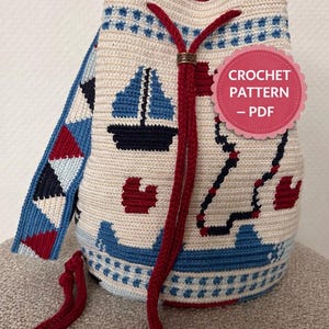 Op de afbeelding: Een crèmekleurige gehaakte emmer tas met een nautisch thema. De tas heeft blauwe zeilboten, rode harten en een blauw-rode band. Het trekkoord is rood en de tas heeft een label met de tekst "CROCHET PATTERN - PDF".