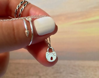 Peut inclure: Un collier en argent avec un petit pendentif en forme de cadenas. Le collier est tenu par une main avec une bague en argent au doigt.
