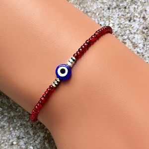 May include: Red beaded bracelet with a blue evil eye charm. The bracelet is made of small red beads and has a silver and gold accent.