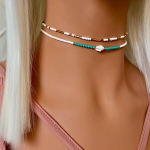 May include: A layered necklace with white and gold beads, a turquoise bead strand, and a small white shell charm.