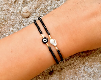 May include: Two black beaded bracelets with a white shell charm and a black evil eye charm. The bracelets are on a person's wrist.