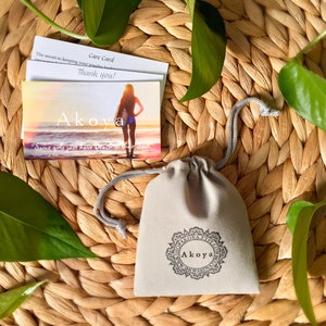 May include: A gray velvet pouch with a drawstring closure. The pouch has a white logo that says "Akoya" with a decorative design. The pouch is sitting on a woven straw surface with green leaves.