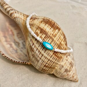 May include: A white beaded bracelet with a turquoise oval charm. The bracelet is lying on a seashell.