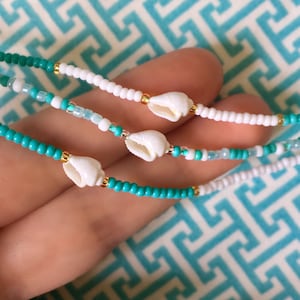 May include: Three turquoise and white beaded bracelets with white shell charms. The bracelets are layered on a hand.