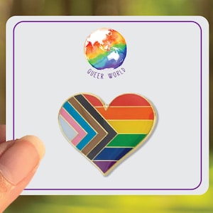 Puede incluir: Un pin dorado en forma de corazón con rayas arcoíris, negras y marrones. El pin dice "Queer World" encima del corazón.