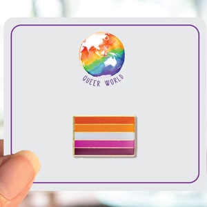 Puede incluir: Una tarjeta blanca con un globo arcoíris y el texto "Queer World" impreso en ella. Un pin dorado con un diseño de bandera del orgullo lésbico está en la tarjeta.
