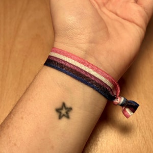 May include: A wrist with a star tattoo and a multi-colored bracelet with pink, white, blue, and black stripes.