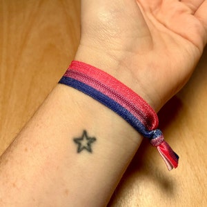 May include: A pink, purple, and blue stretchy bracelet with a knot on the end. The bracelet is worn on a person's wrist, and a small star tattoo is visible on the wrist.