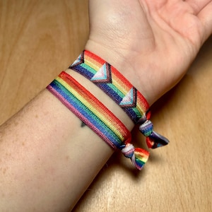 May include: Two colorful bracelets on a person's wrist. The top bracelet is a rainbow pattern with a white triangle design. The bottom bracelet is a rainbow pattern with a white triangle design.