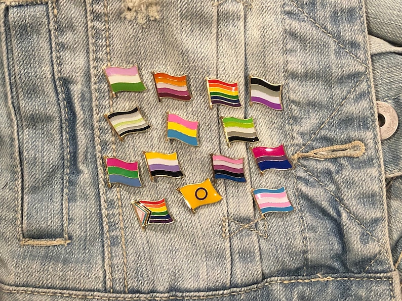 May include: A collection of enamel pins featuring various pride flags, including the transgender, lesbian, gay, bisexual, pansexual, asexual, and intersex flags. The pins are arranged on a denim background.