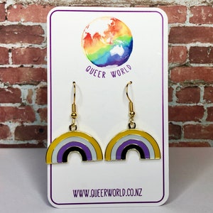 May include: A pair of gold earrings with a rainbow design. The rainbow is in the colors of the ace flag: yellow, white, purple, black, and white. The earrings are on a white card with the text "Queer World" and "www.queerworld.co.nz".