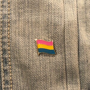 May include: A gold-colored pin with a pansexual pride flag design. The flag has three horizontal stripes: pink, yellow, and blue.