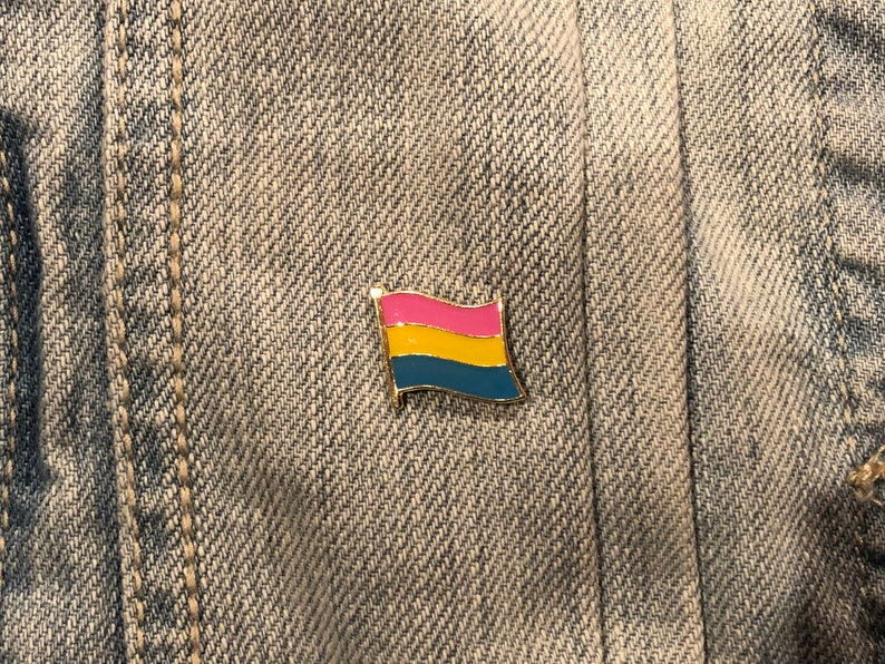 May include: A gold-colored pin with a pansexual pride flag design. The flag has three horizontal stripes: pink, yellow, and blue.