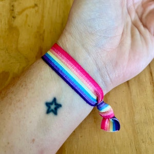 May include: A rainbow coloured elastic bracelet with a knot on the wrist. A small black star tattoo is visible on the wrist.