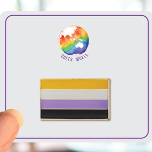 May include: A gold-colored pin with a horizontal flag design. The flag has five stripes: yellow, white, light purple, black, and yellow. The pin features the text "Queer World" and a rainbow globe.
