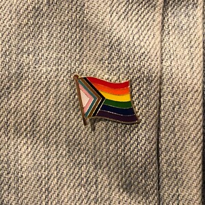 May include: A gold-colored pin with a rainbow flag design. The flag has a white triangle with a pink, blue, and light blue stripe, followed by a black and brown stripe, and then the traditional rainbow colors of red, orange, yellow, green, blue, indigo, and violet.