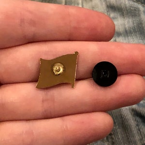 May include: Gold-colored metal pin with a flag design. The back of the pin has a black rubber backing with a small metal post.